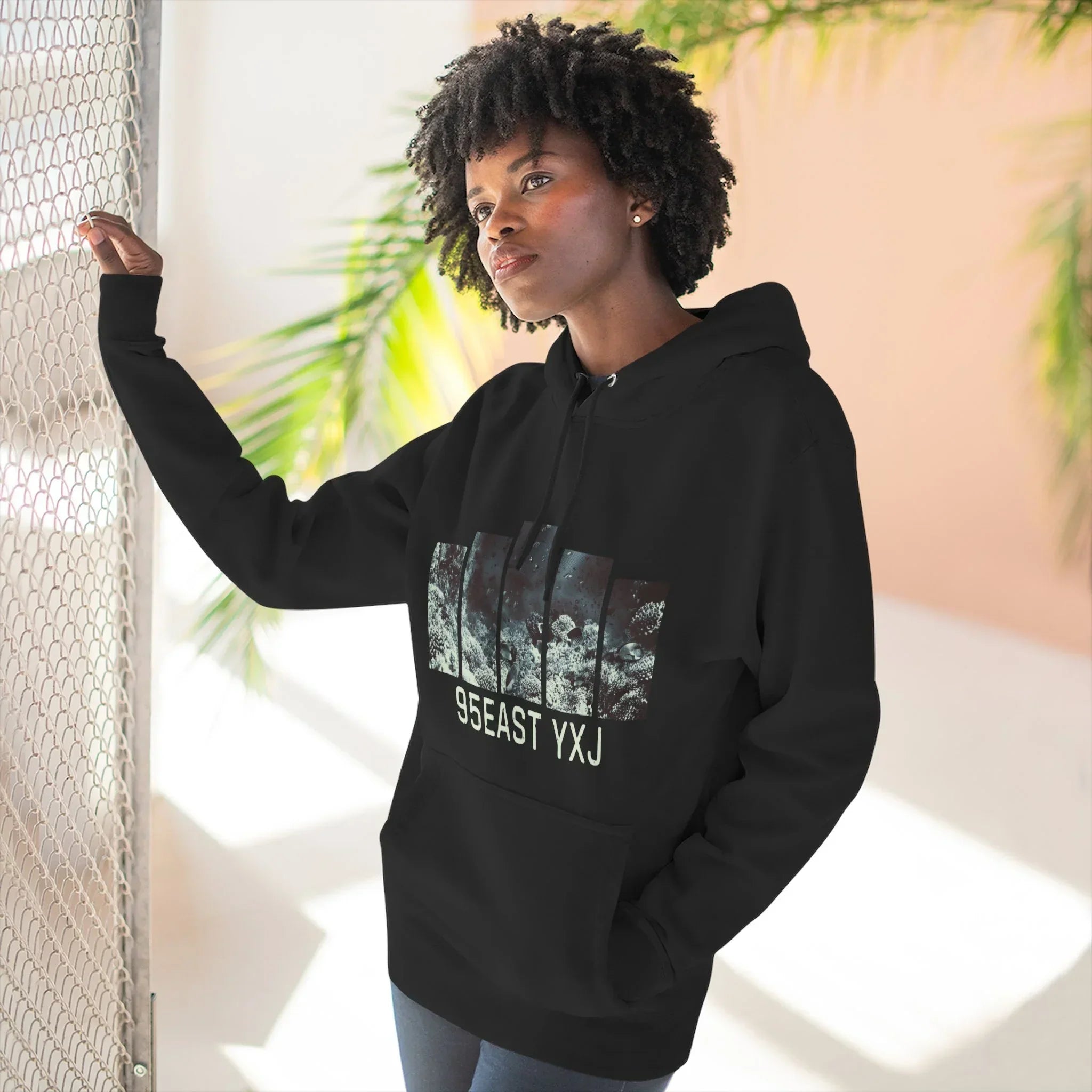 Woman wearing a black hoodie with graphic design and 95EAST YXJ text, standing by a fence