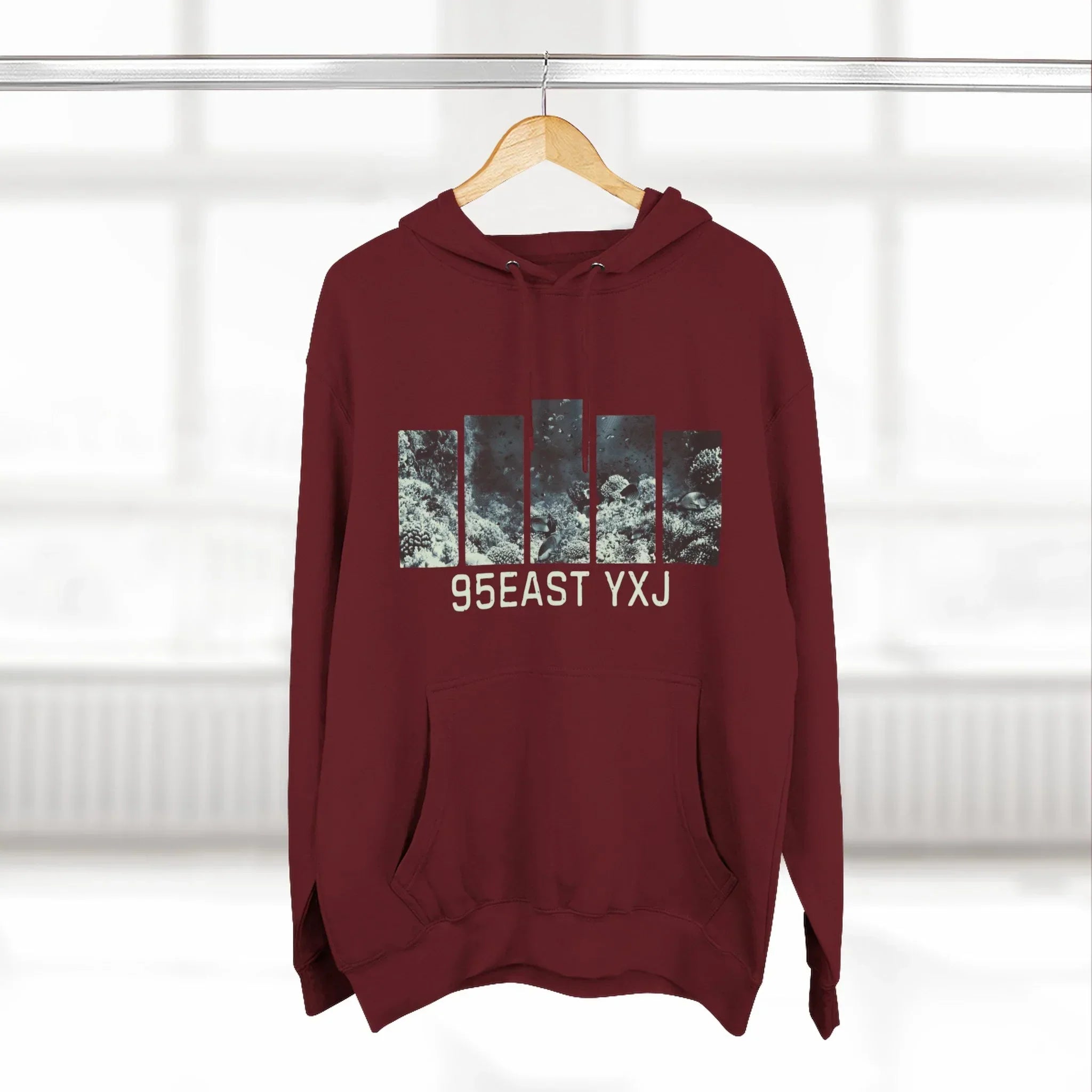 Maroon hoodie with graphic print and 95EAST YXJ text on hanger in bright room