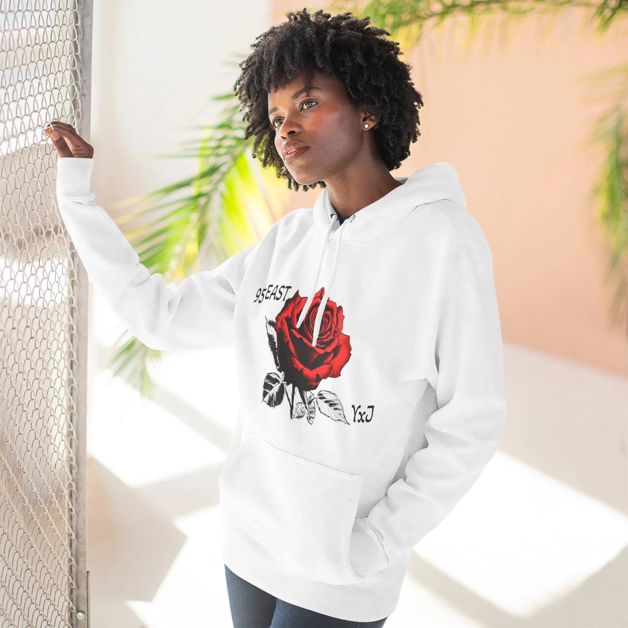White hoodie with red rose graphic and '95EAST YxJ' text, worn by woman near mesh fence
