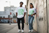 Man and woman wearing white graphic tees with green text and silhouette, standing near graffiti wall