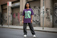 Purple graphic tee with bold green text and silhouette, paired with black jeans and white sneakers on urban street.
