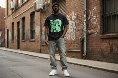 Man wearing black t-shirt with green graphic and text, standing on urban street with brick walls.