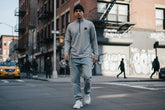 Man in gray quarter-zip sweatshirt and ripped jeans walking on urban street, city fashion