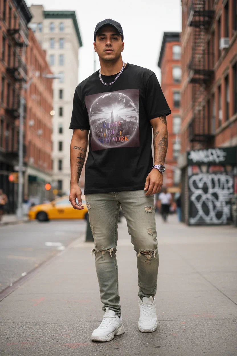 Man in black graphic tee with NYC skyline print, ripped jeans, white sneakers, urban street backdrop
