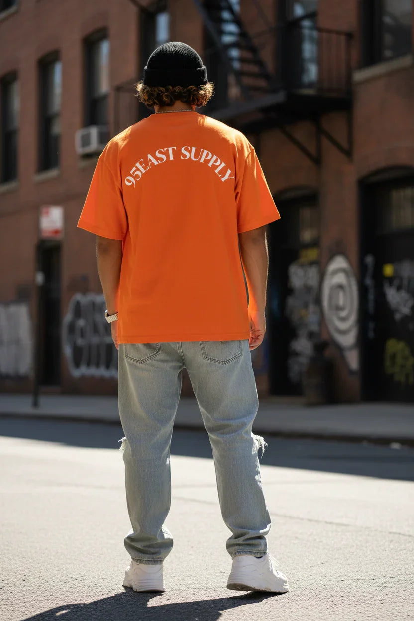 Man wearing orange 95EAST SUPPLY tee, light jeans, and beanie on urban street