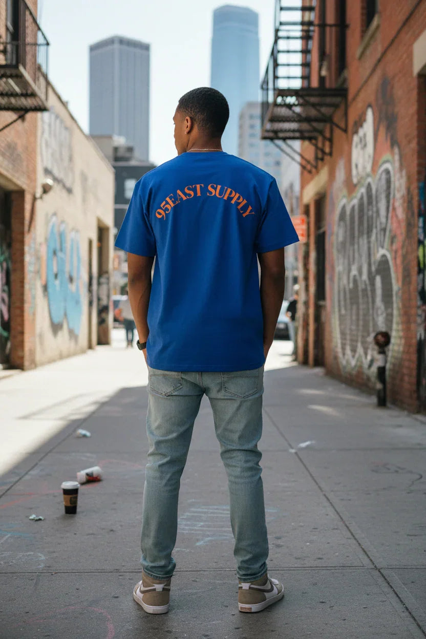 Man wearing blue 95EAST SUPPLY tee in urban alley, graffiti walls, city skyline backdrop