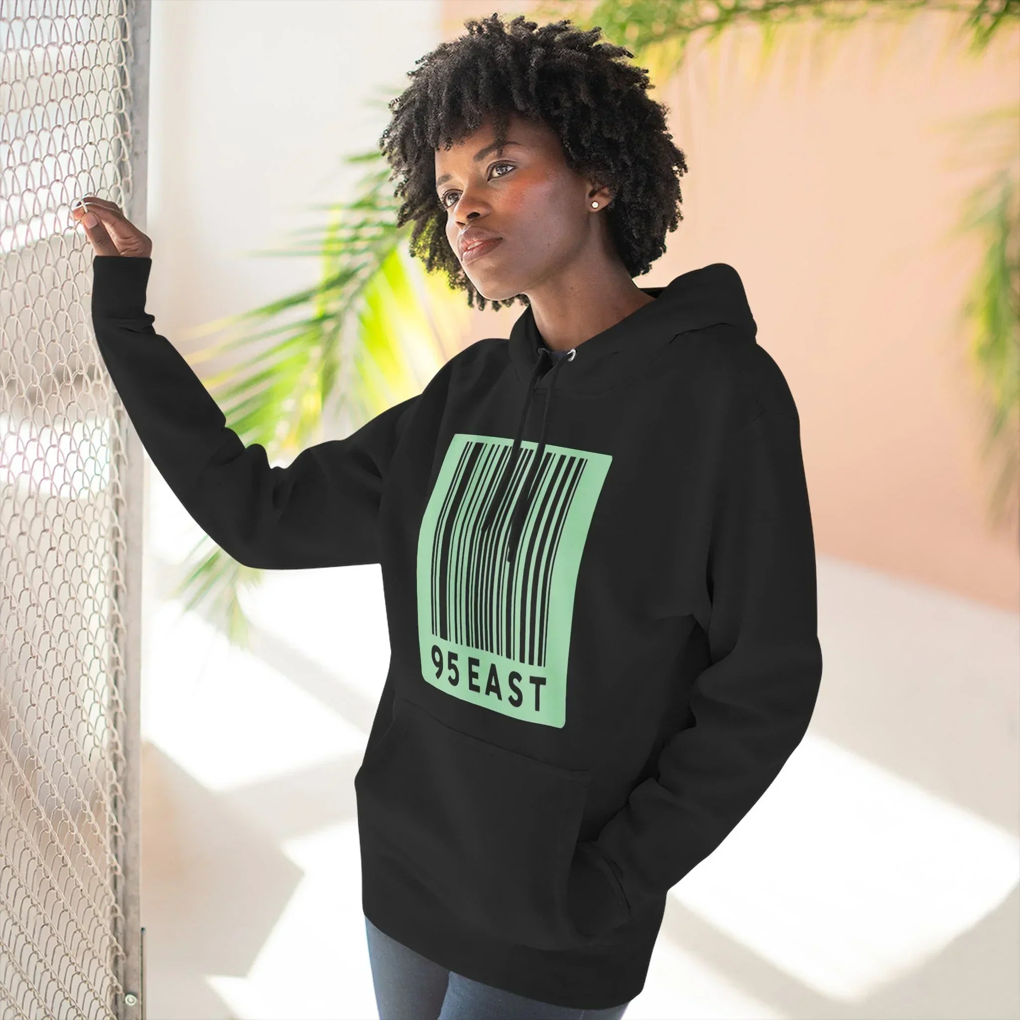 Black hoodie with green barcode design and 95 EAST text, worn by woman near mesh fence and plants