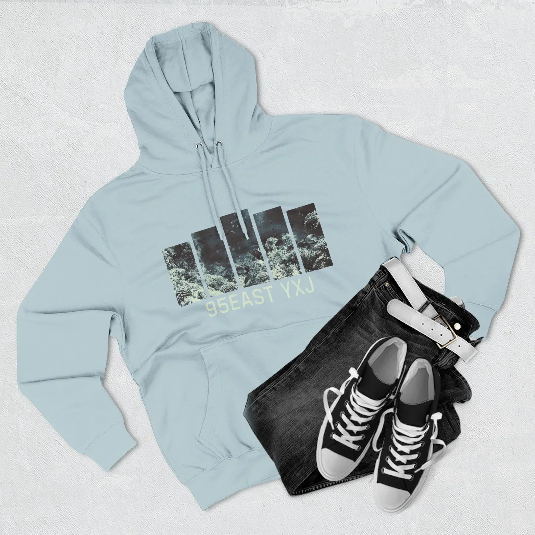 Flat lay of light blue 95EAST YXJ graphic hoodie, black jeans, white belt, and black sneakers.