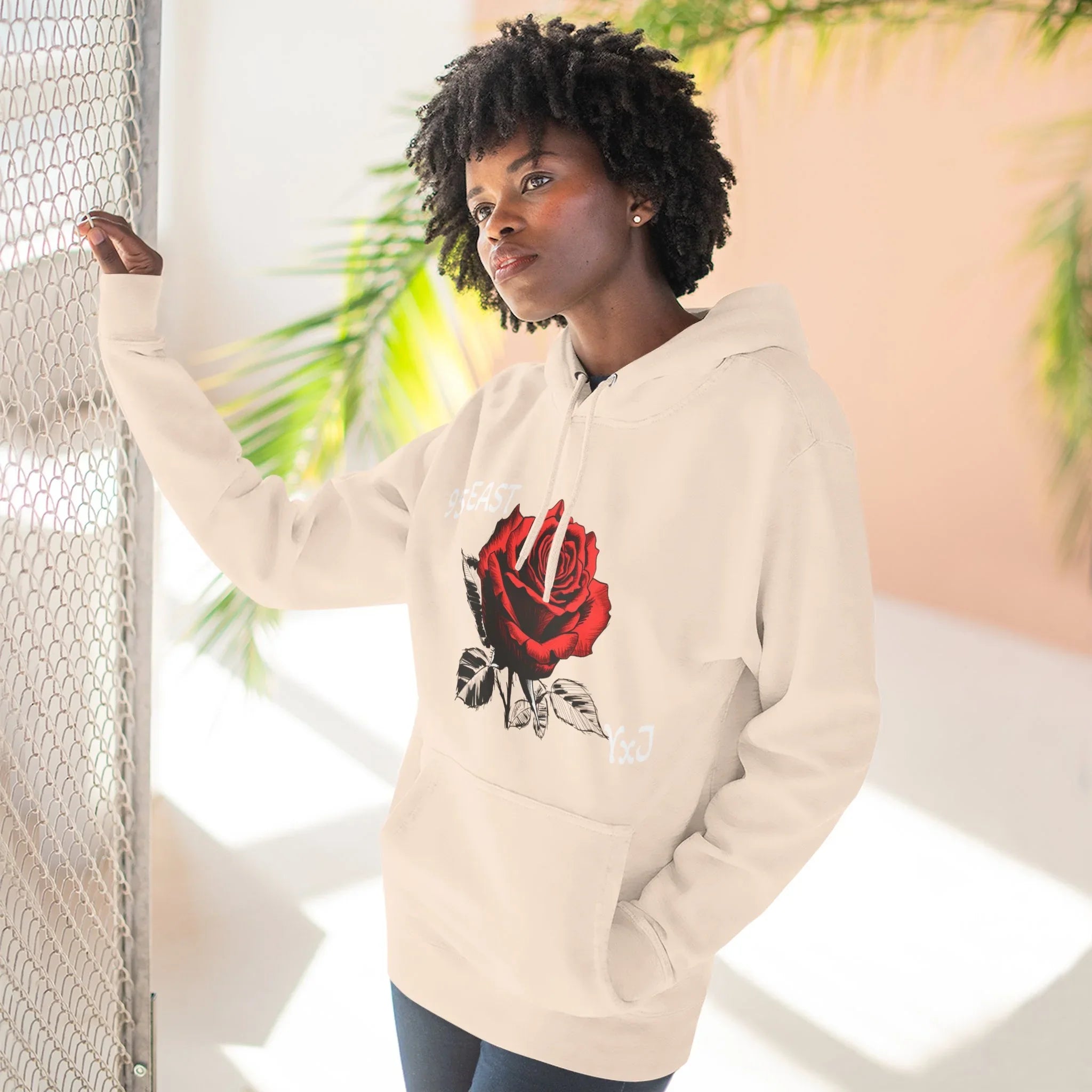 Cream hoodie with red rose graphic and YxJ text, worn by woman near mesh fence
