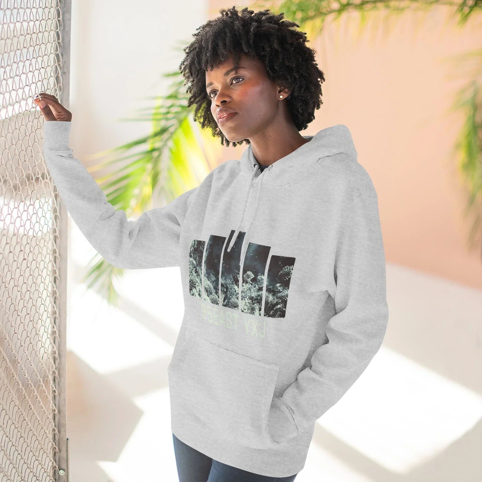 Woman wearing a light gray graphic hoodie with bold black print, standing by a fence indoors