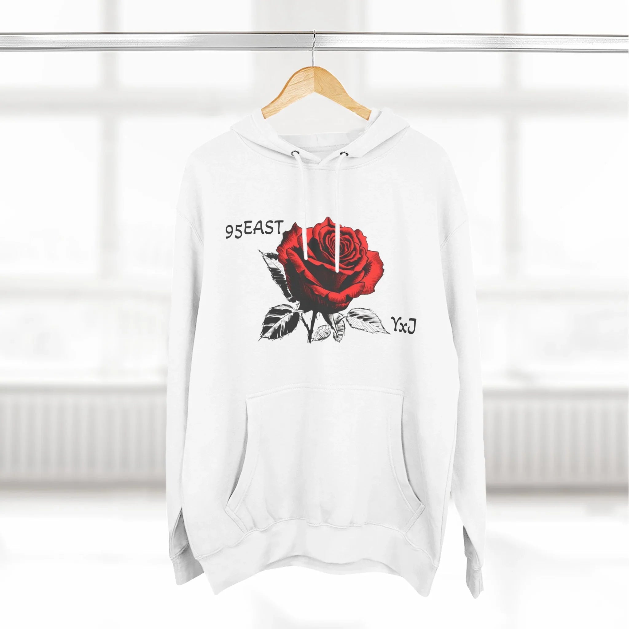 White hoodie with red rose graphic and 95EAST YxJ text, hanging on wood hanger