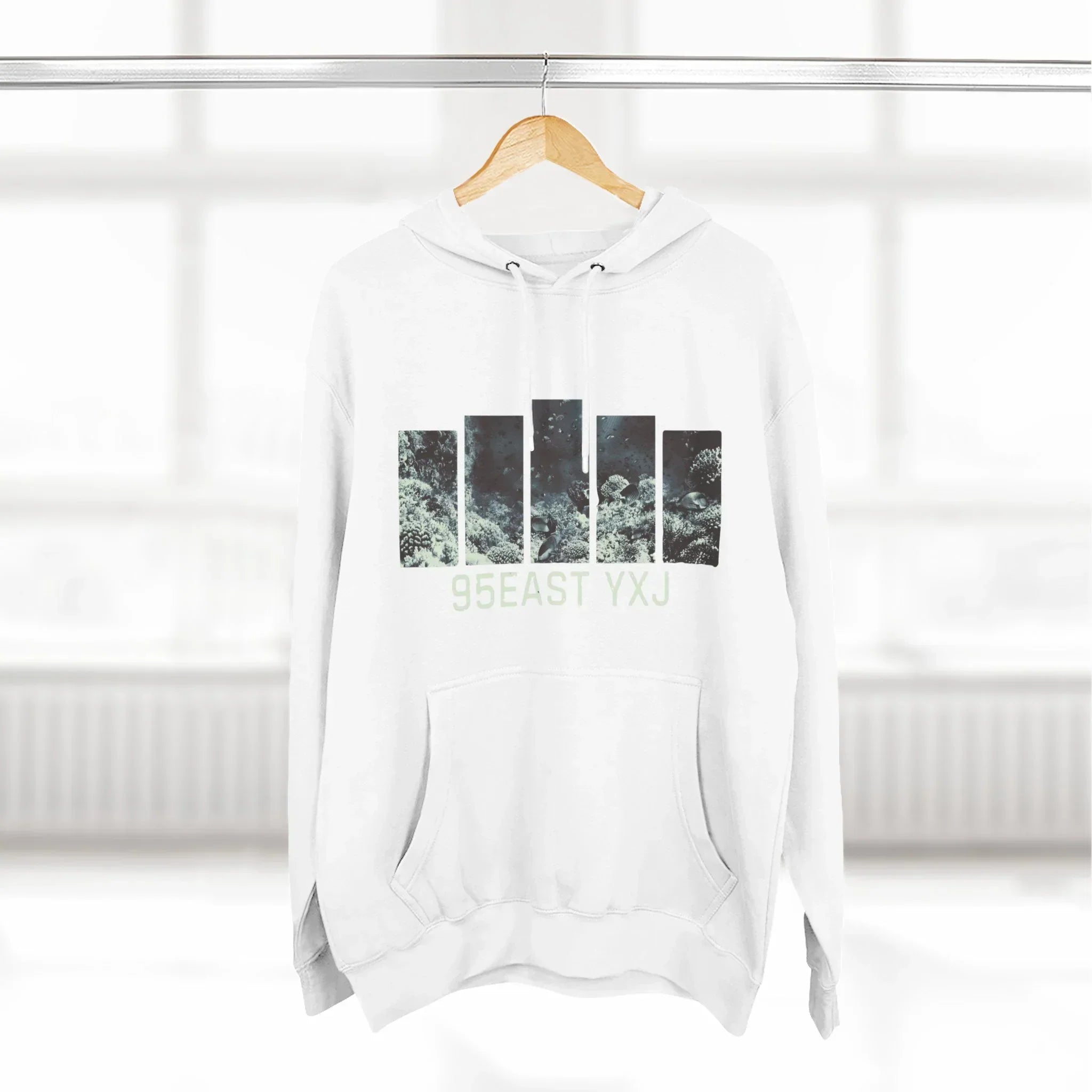 White hoodie with graphic nature print and 95EAST YXJ text on hanger, bright room background