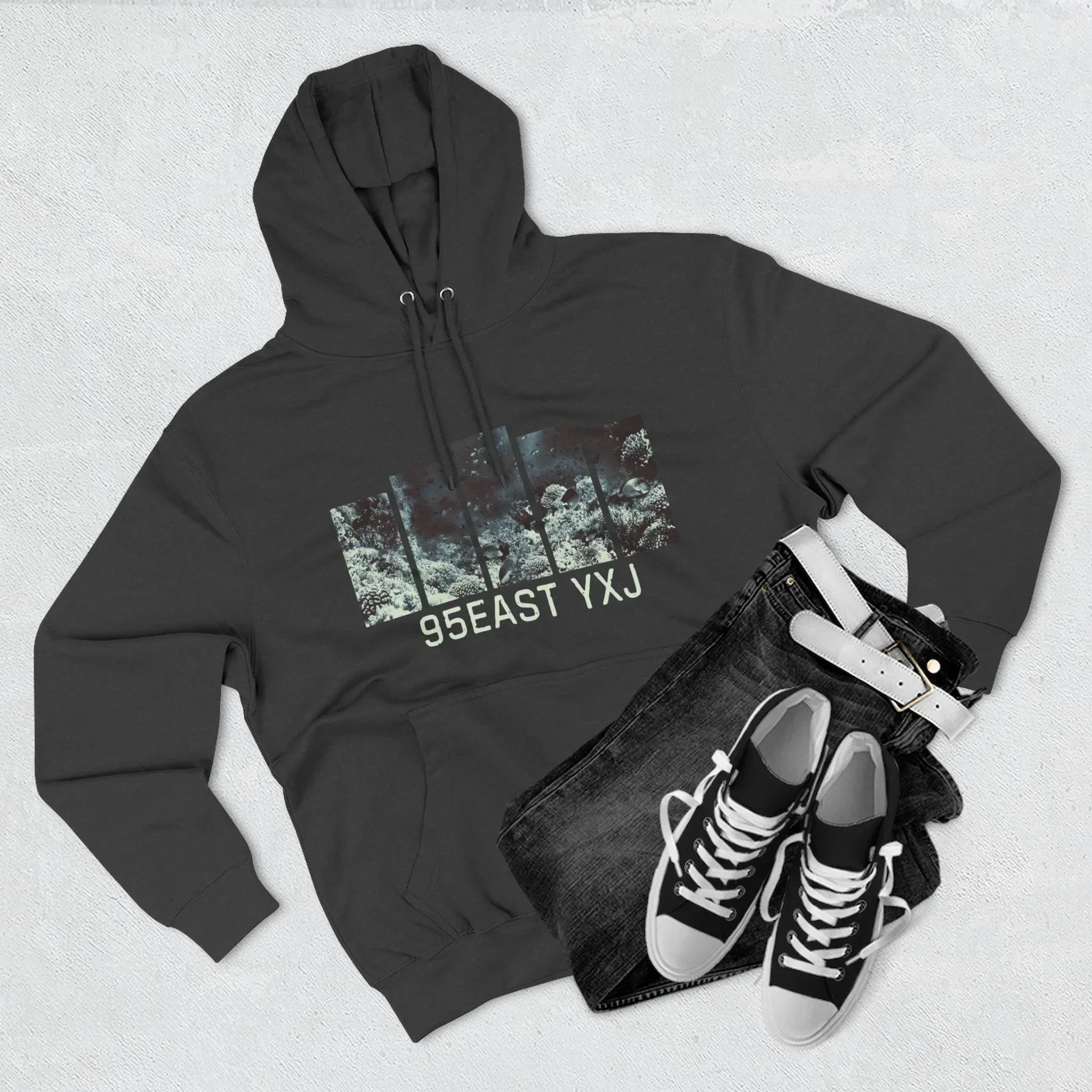 Black hoodie with abstract graphic and 95EAST YXJ text, paired with black jeans, white belt, and sneakers