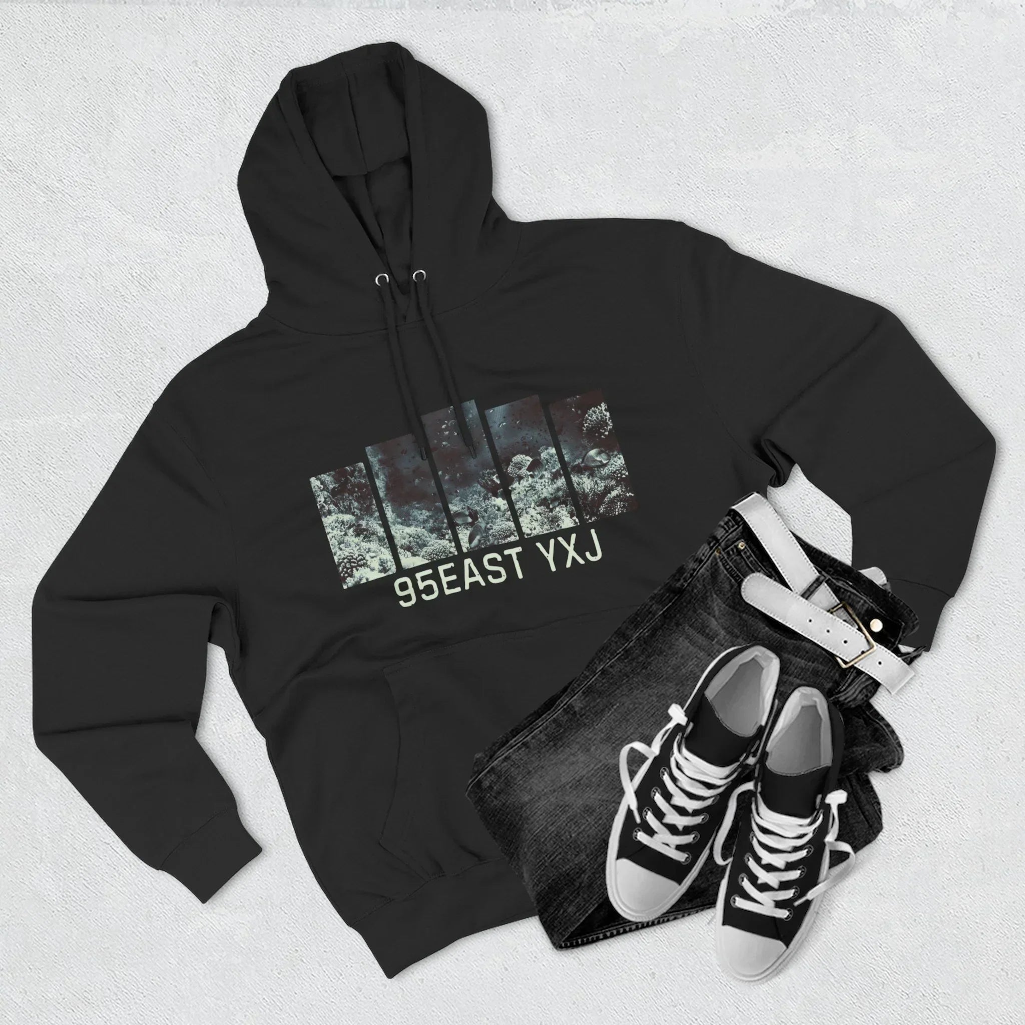 Flat lay of black graphic hoodie with 95EAST YXJ print, black jeans, belt, and sneakers