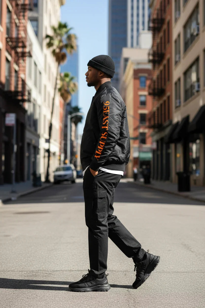 Man in black bomber jacket with orange text, walking urban street, modern East Coast style