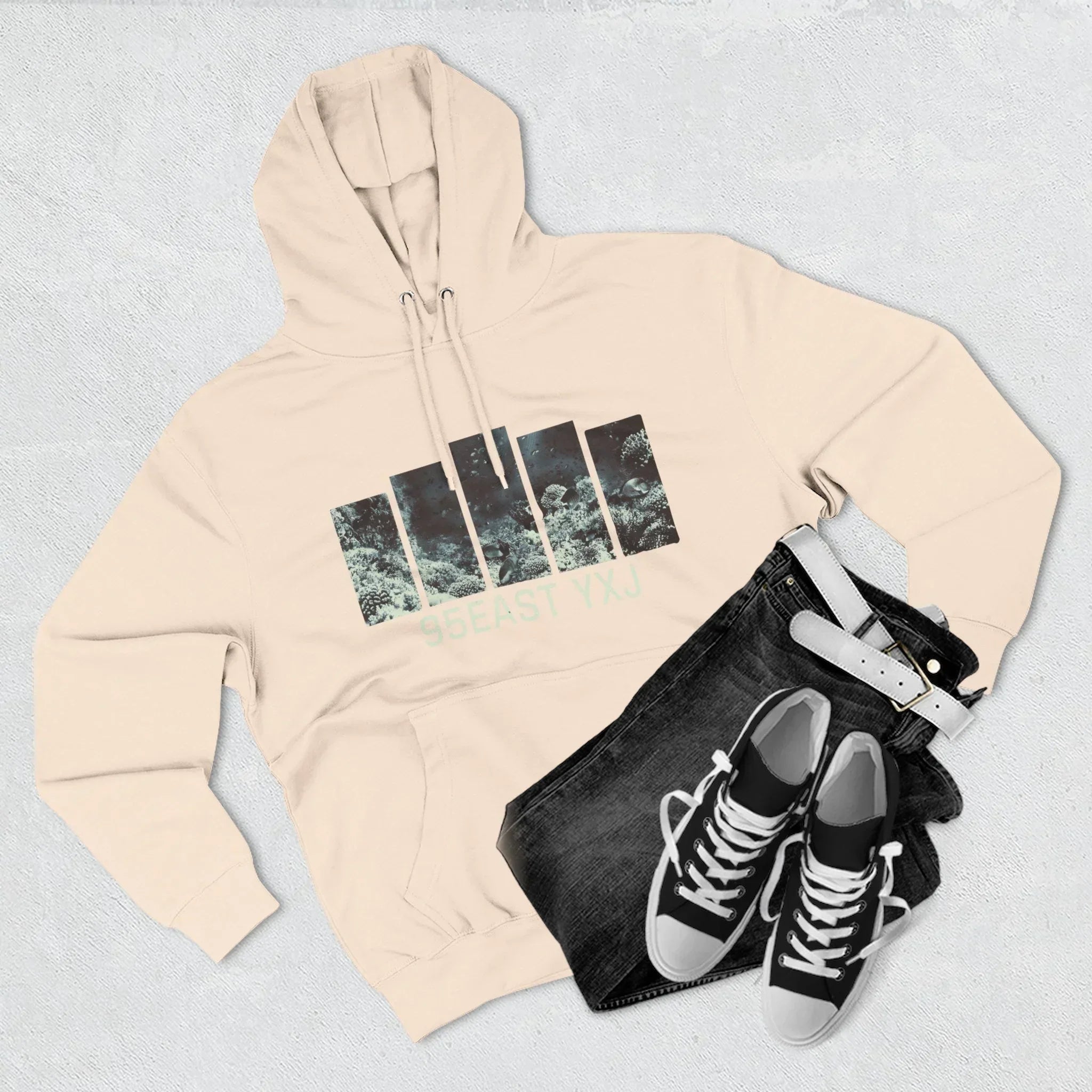 Beige hoodie with graphic print and 95EAST YXJ text, paired with black jeans, sneakers, and white belt.
