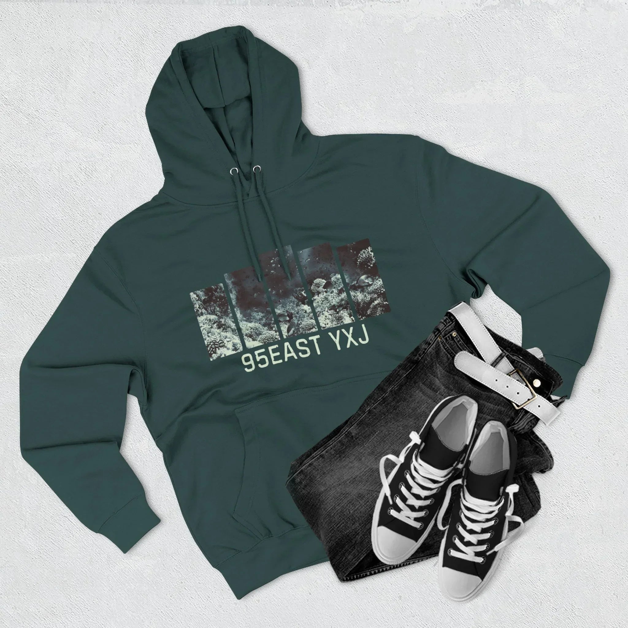 Dark green hoodie with 95EAST YXJ graphic, black jeans, white belt, and black sneakers laid out on light background.