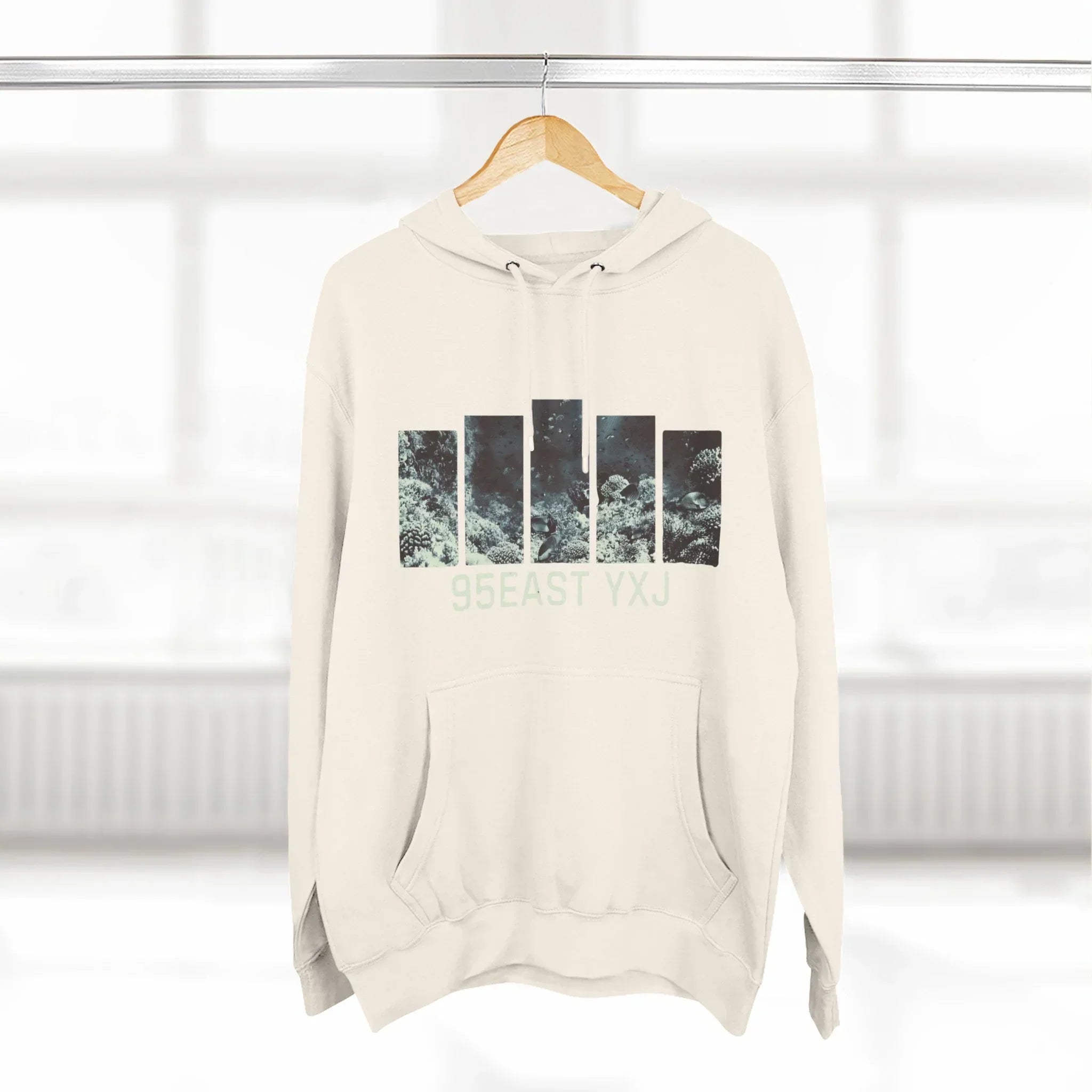 Beige hoodie with abstract black print and '95EAST YXJ' text, hanging on wooden hanger.