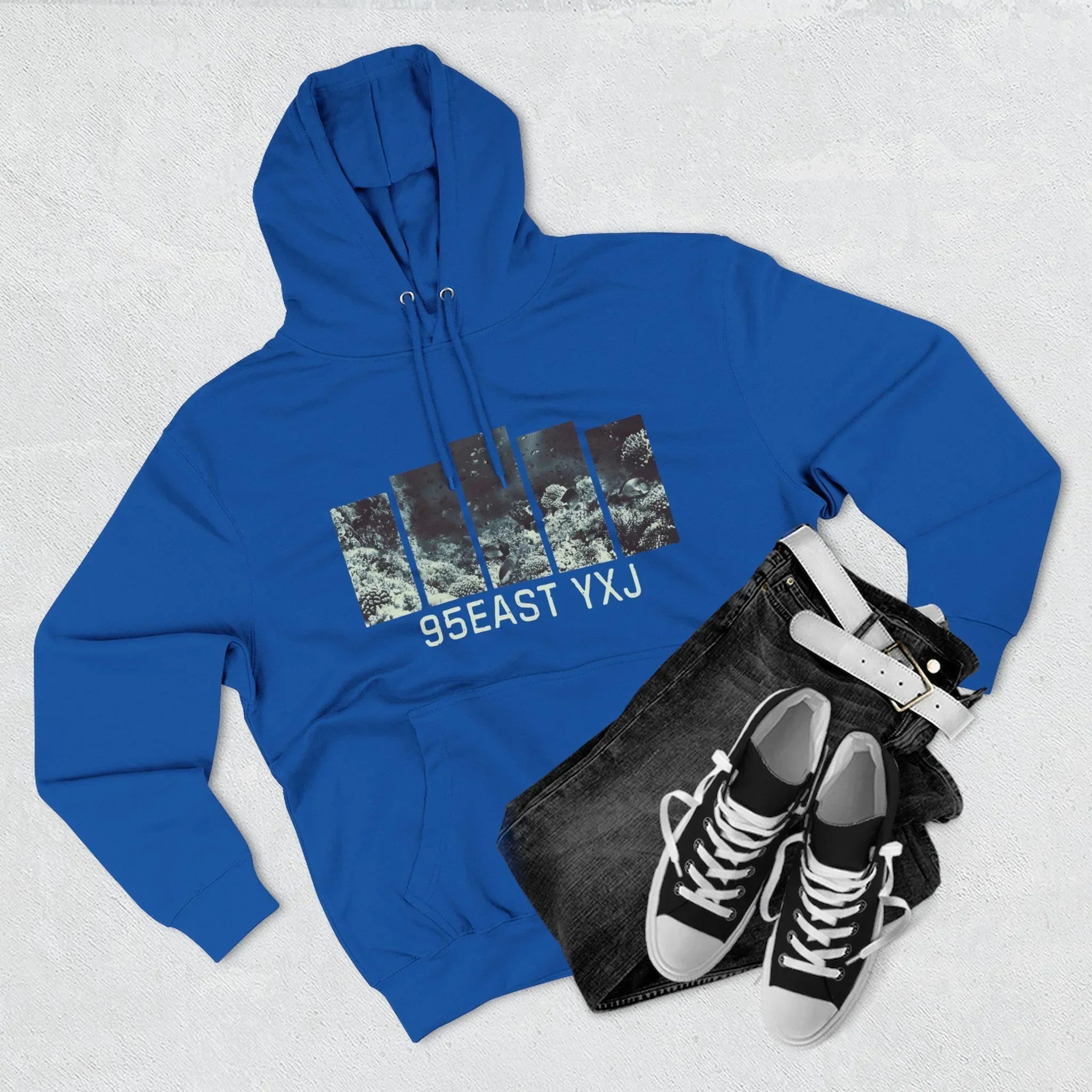 Blue 95EAST YXJ hoodie with graphic print, black jeans, white belt, and black sneakers.