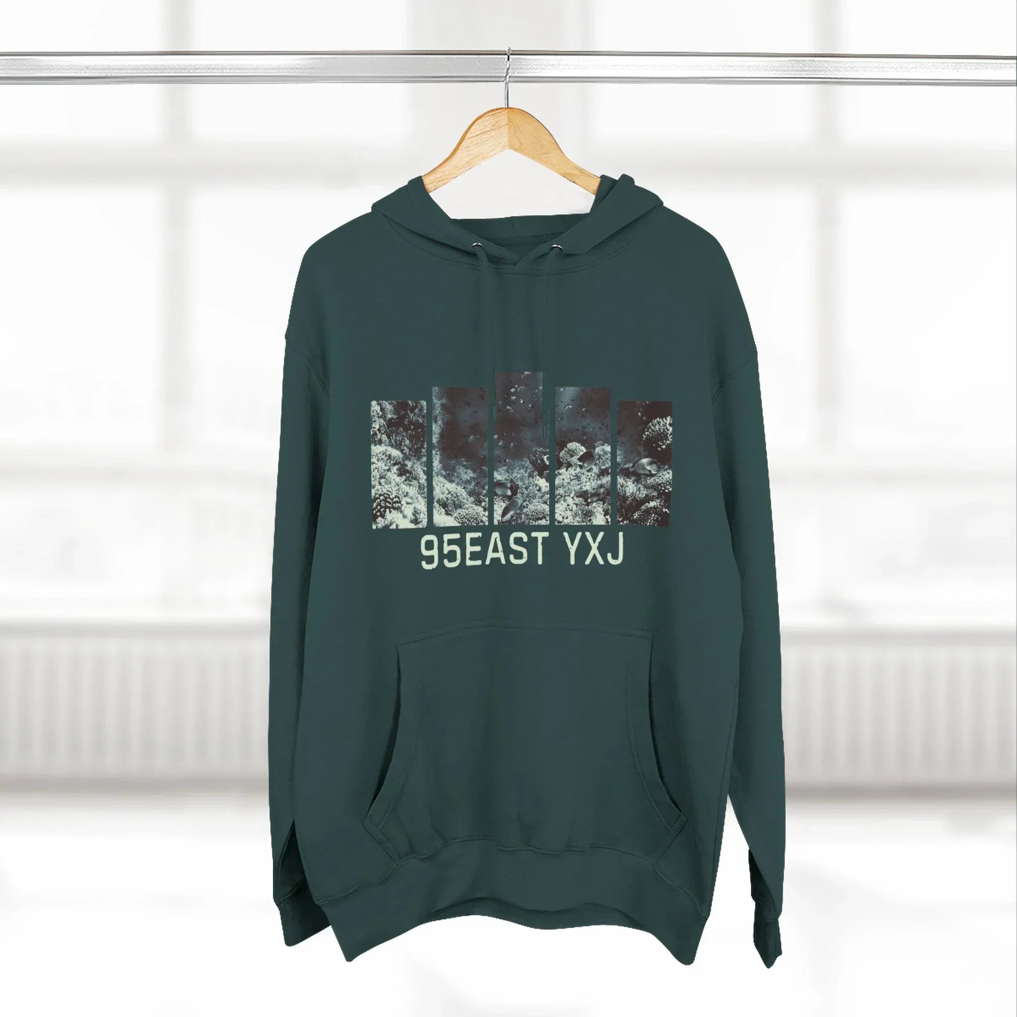 Dark green hoodie with abstract graphic and 95EAST YXJ text, hanging on wooden hanger