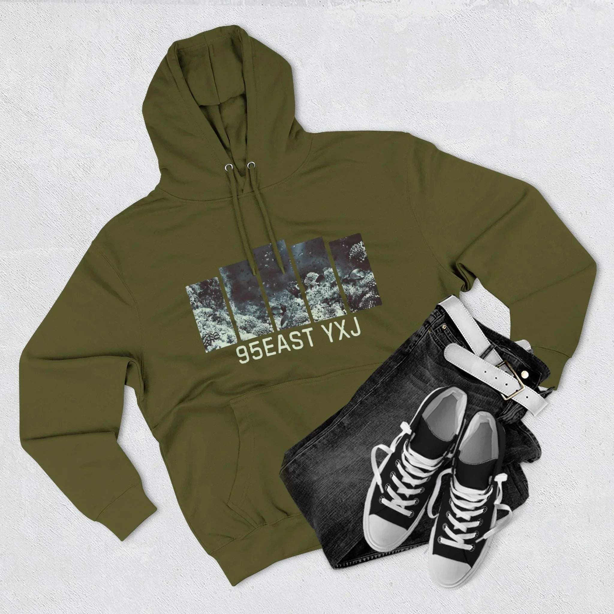 Olive green hoodie with graphic print and '95EAST YXJ' text, paired with black jeans, white belt, and black sneakers on a light background.