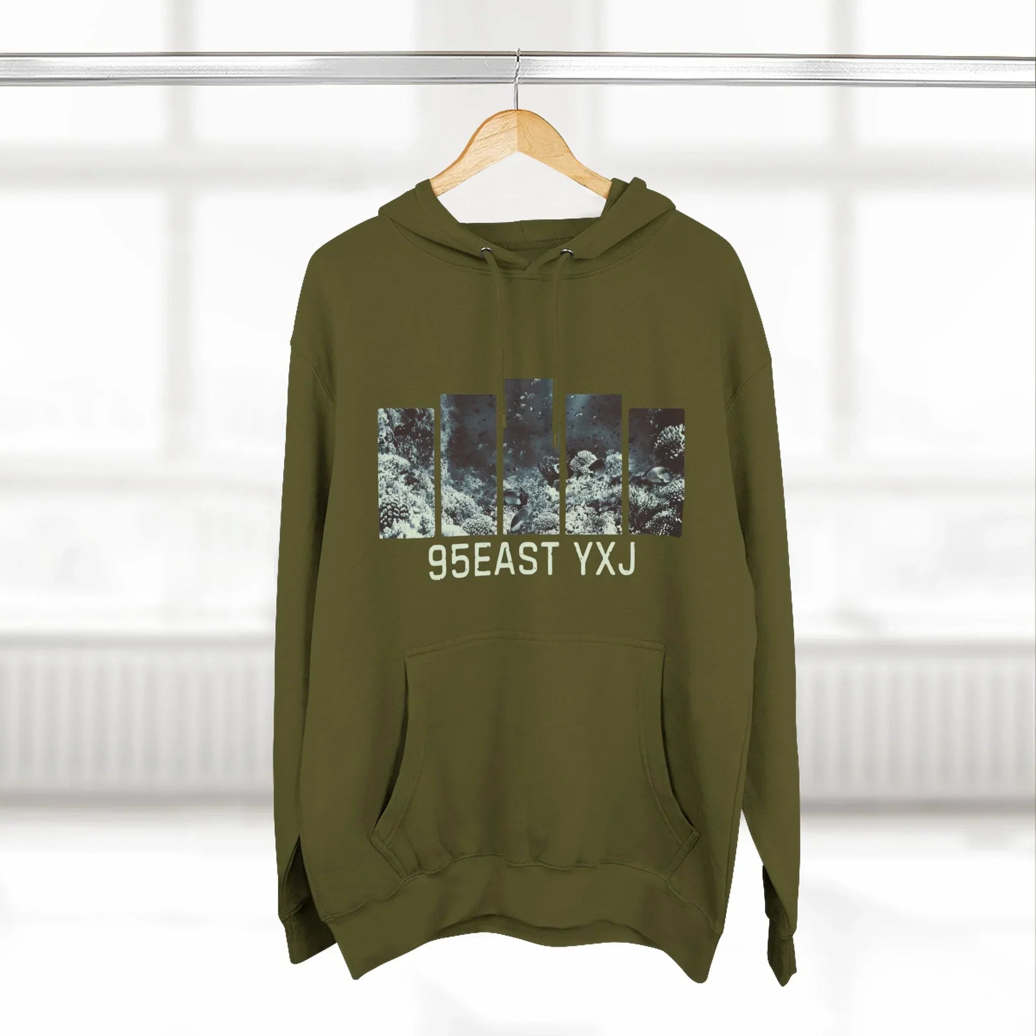 Olive green hoodie with abstract graphic and '95EAST YXJ' text, hanging on a wooden hanger