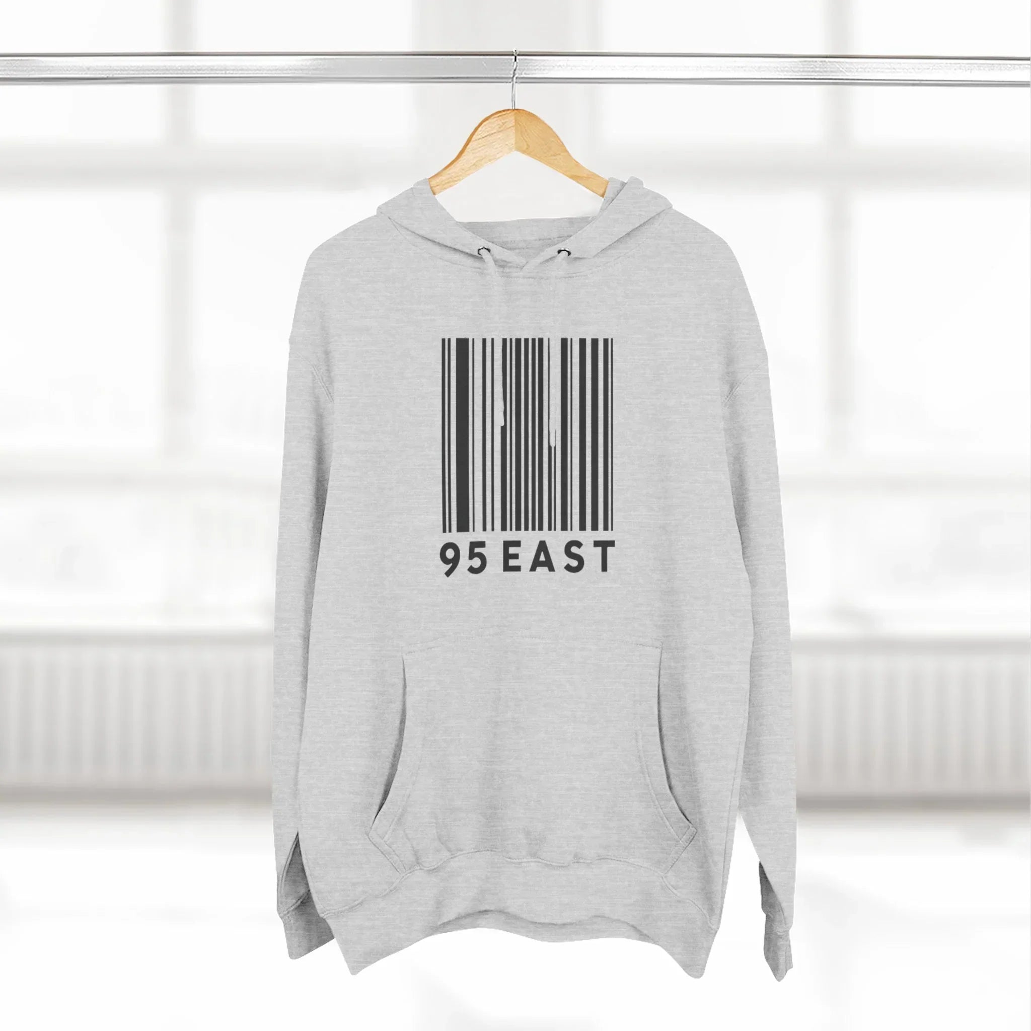 Light gray hoodie with black barcode and '95 EAST' text on hanger in bright room